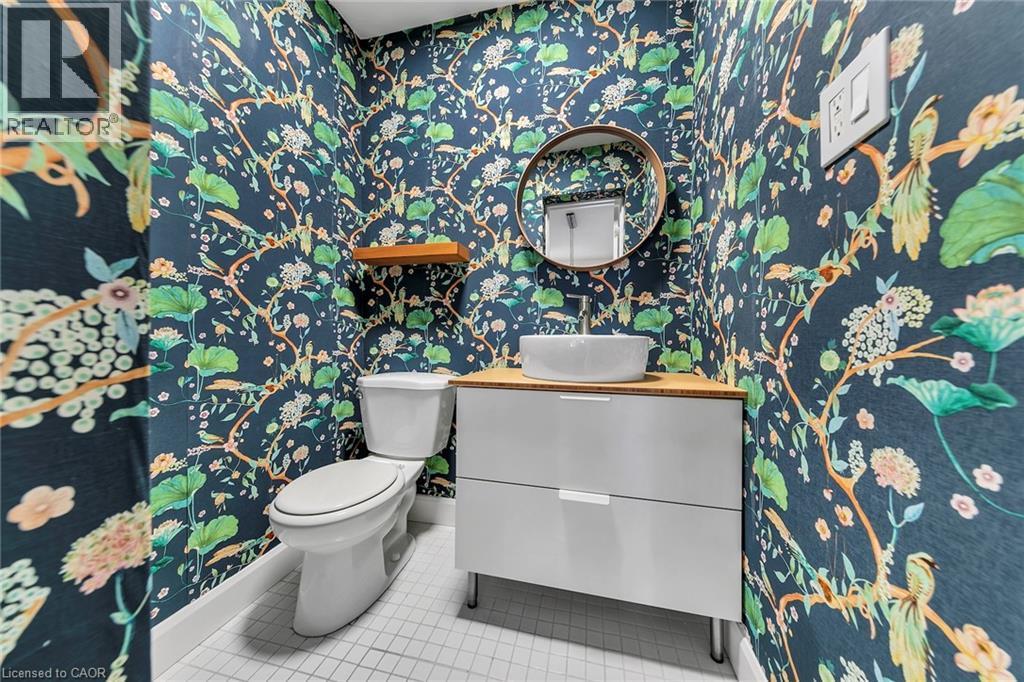 Powder room featuring botanical wallpaper, a white vessel sink, a light wood-finish countertop, a round frameless mirror, and white square floor tiles - 501 Ferguson Avenue N, Hamilton, ON - Indoor Photo Showing Bathroom
