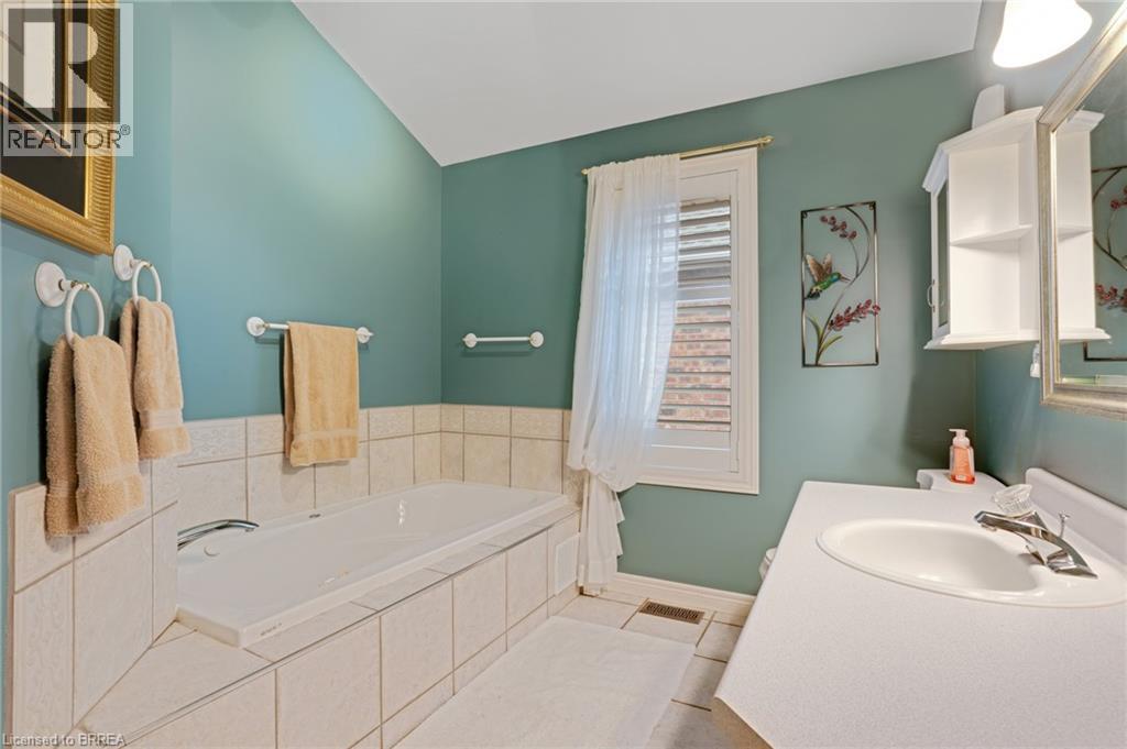Spacious bathroom featuring a built-in bathtub with tiled surround, a vanity with an integrated sink, a wall-mounted medicine cabinet, and a window with white louvered shutters - 422 Powerline Road Unit# 10, Brantford, ON - Indoor Photo Showing Bathroom