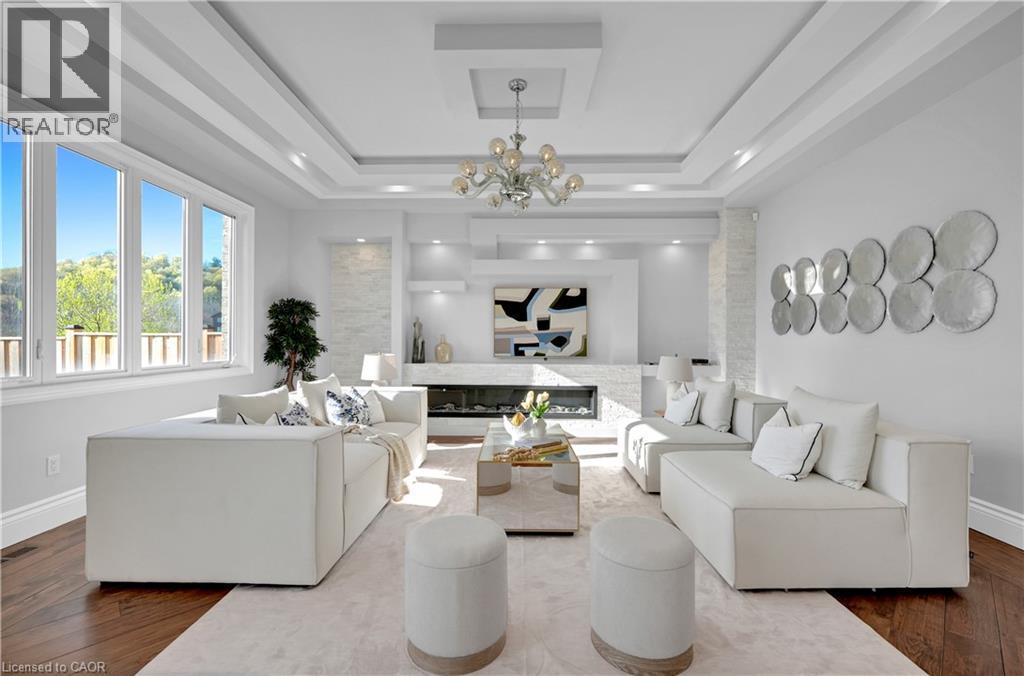 Spacious living area featuring expansive windows, wood-finish flooring, a contemporary fireplace with stone surround, recessed lighting, and a multi-tiered tray ceiling with a chandelier - 7 Riesling Court, Hamilton, ON - Indoor Photo Showing Living Room