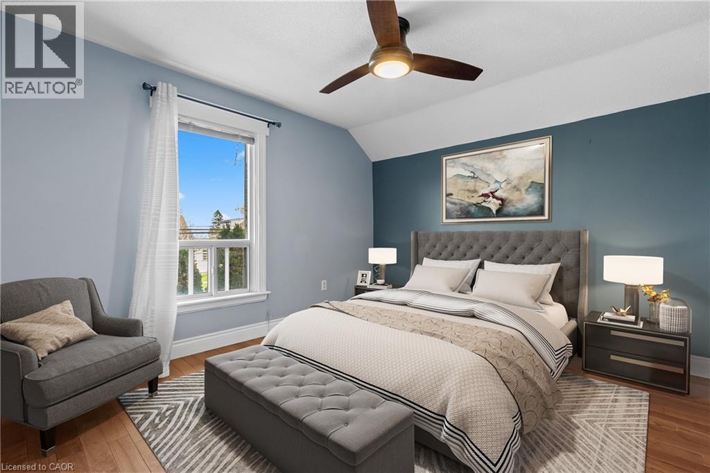 Wood-finish flooring with a multi-tone area rug, featuring a large window with white trim, a ceiling fan with integrated lighting, and a prominent accent wall (Virtually Staged) - 204 Beach Boulevard, Hamilton, ON - Indoor Photo Showing Bedroom