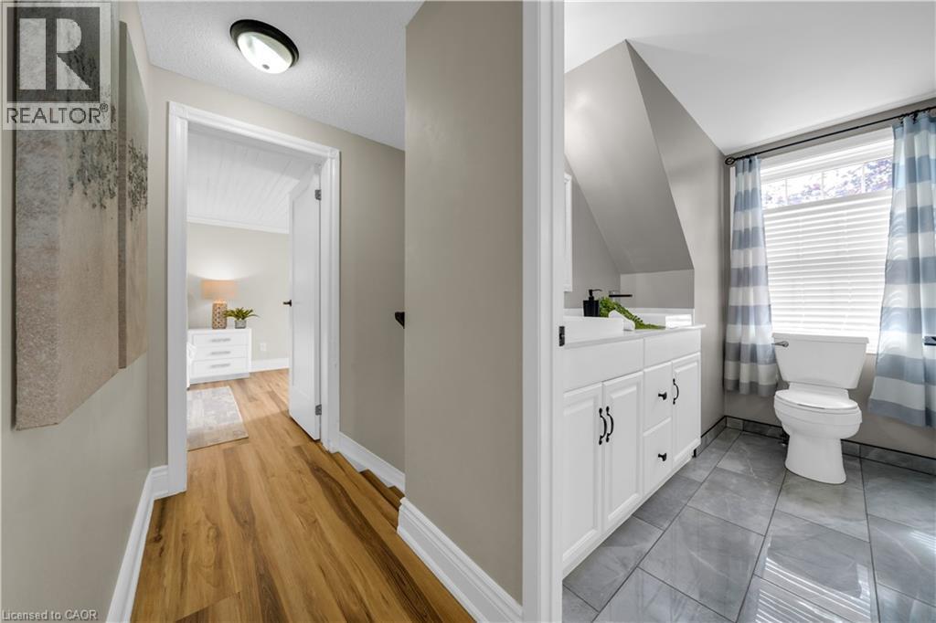 Hallway featuring wood-finish flooring, leading to a bathroom with a white vanity, a toilet, and a window with blinds - 26 Peachtree Crescent, Cambridge, ON - Indoor Photo Showing Bathroom
