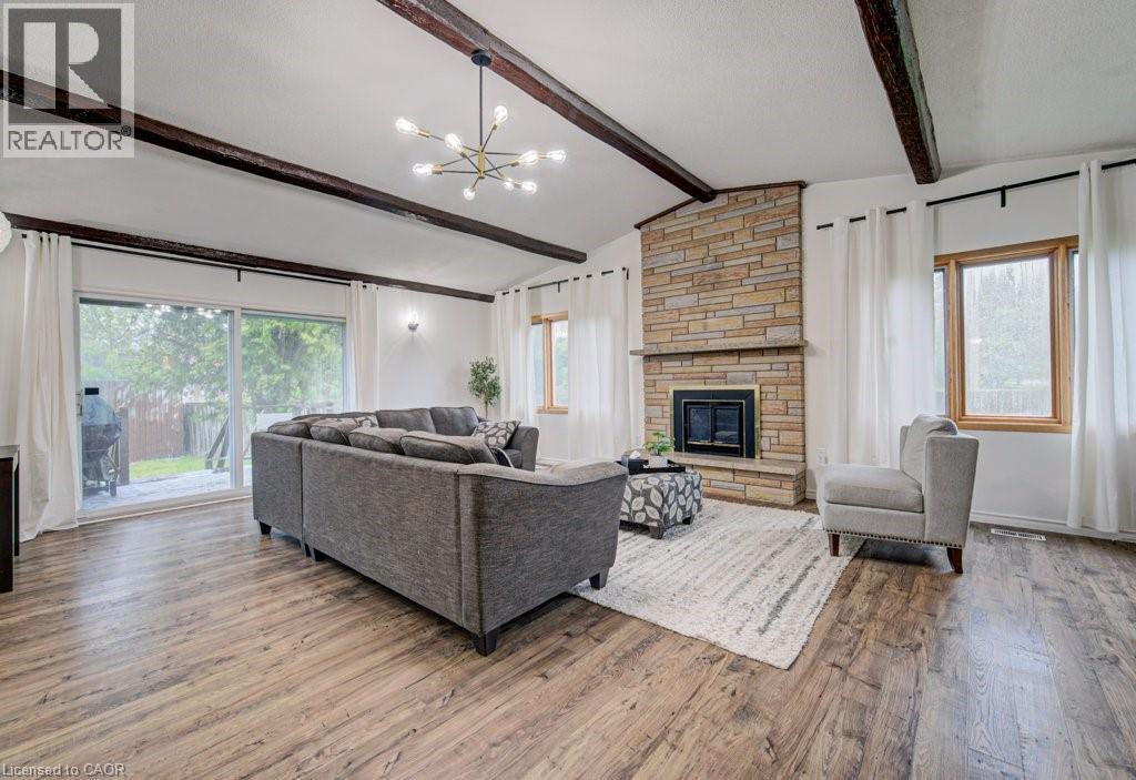 Spacious living area featuring wood-finish flooring, a stone-clad fireplace, exposed wood beams, and a modern chandelier - 128 Pollock Avenue, Cambridge, ON - Indoor Photo Showing Living Room With Fireplace