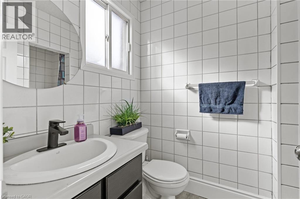 Bathroom featuring white square tile walls, a window with privacy glass, a vanity with an integrated sink, a matte black faucet, and a wall-mounted round mirror - 27 Barbara Court, Hamilton, ON - Indoor Photo Showing Bathroom