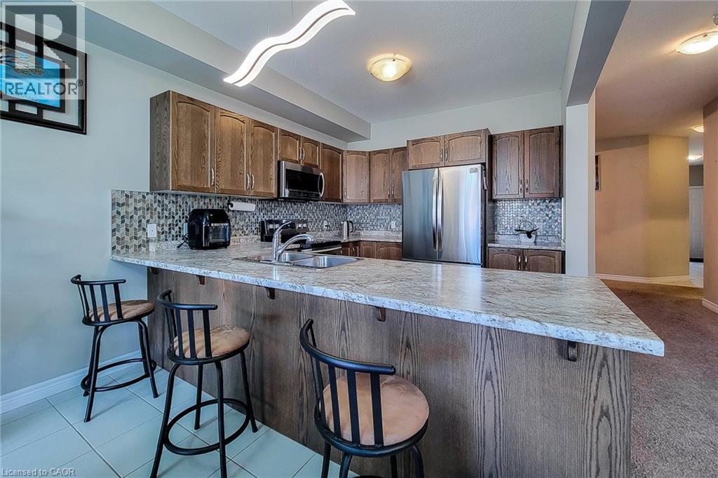 Kitchen featuring wood-finish cabinetry, stainless steel appliances, a multi-tone tile backsplash, light-toned countertops, and a breakfast bar with seating - 40 Fairgrounds Drive, Binbrook, ON - Indoor Photo Showing Kitchen With Double Sink