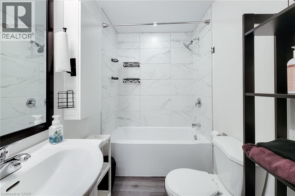 Bathroom featuring a white vanity with a single basin sink, a framed mirror, a built-in bathtub with shower, white tiled walls, and wood-finish flooring - 40 Fairgrounds Drive, Binbrook, ON - Indoor Photo Showing Bathroom