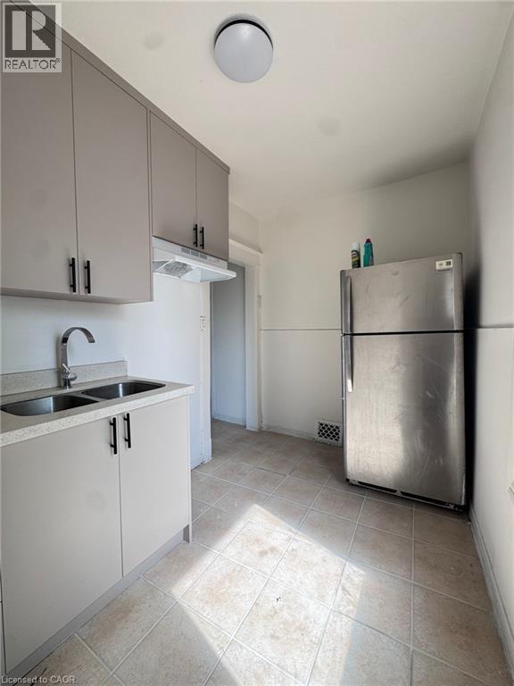 Kitchen featuring gray cabinetry with matte black hardware, a double basin stainless steel sink, a stainless steel refrigerator, and tiled flooring - 10 Fairleigh Avenue N, Hamilton, ON - Indoor Photo Showing Kitchen With Double Sink