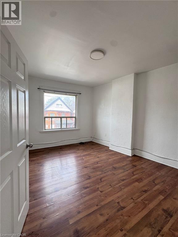 Room featuring wood-finish flooring, white painted walls, a window with exterior residential structures visible, a white paneled door, and a flush-mount ceiling light fixture - 10 Fairleigh Avenue N, Hamilton, ON - Indoor Photo Showing Other Room