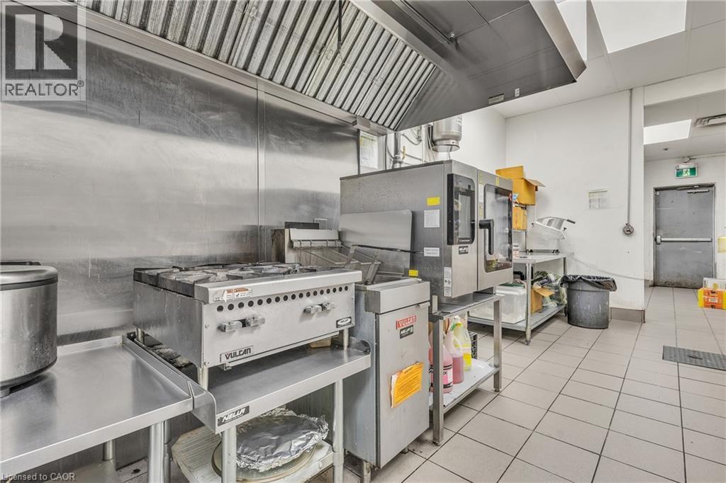 Industrial kitchen featuring stainless steel countertops, a commercial range with overhead ventilation, and tiled flooring - 5 Manitou Drive Unit# 106B, Kitchener, ON