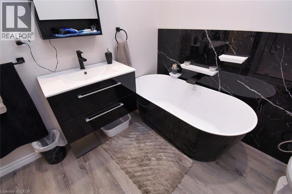 Bathroom featuring a freestanding black and white soaking tub, a wall-mounted vanity with integrated sink and black cabinetry, black accent wall with white veining, wood-finish flooring, and matte bla - 5 Ralph Street, Turkey Point, ON - Indoor Photo Showing Bathroom