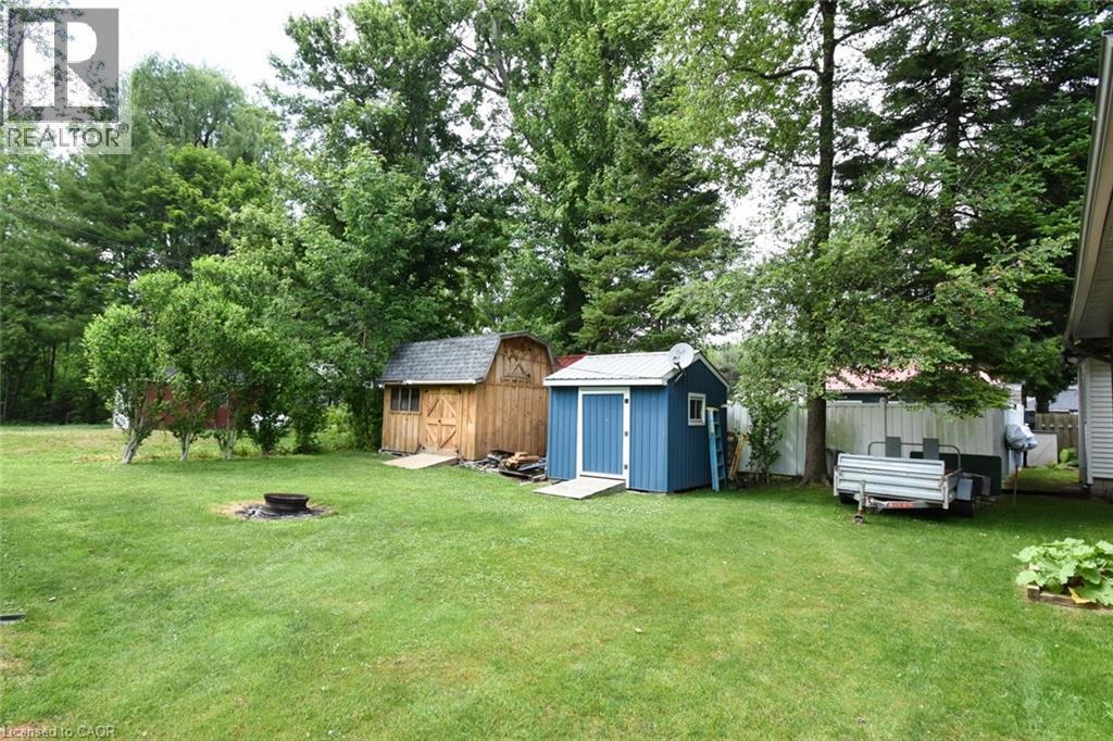 Expansive green lawn with established trees, featuring a natural wood-finish storage shed with barn-style doors, a blue storage shed with a white roof, and a built-in fire pit - 5 Ralph Street, Turkey Point, ON - Outdoor With Backyard