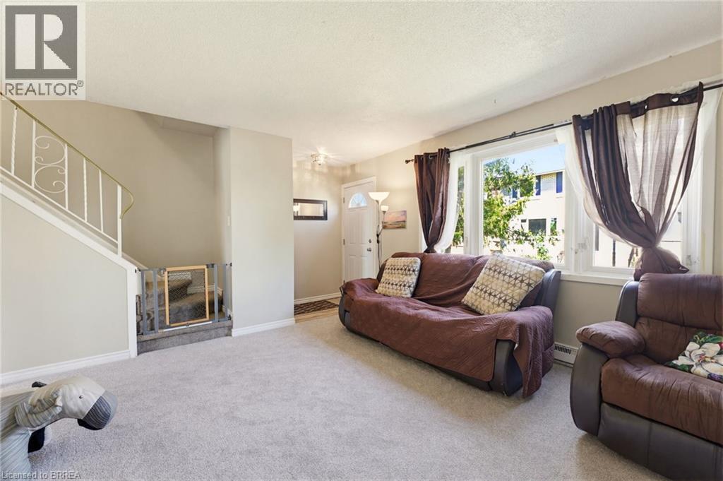 Carpeted living area featuring a large window, light beige wall paint, white trim, and a white front entry door with a half-moon window - 26 Coachwood Road Unit# 6, Brantford, ON - Indoor Photo Showing Living Room
