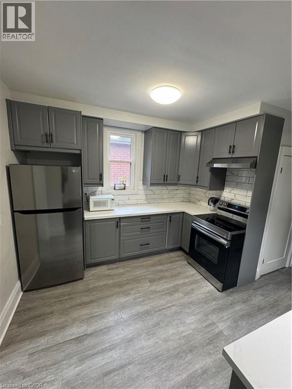 Modern kitchen featuring gray shaker cabinetry, light countertops, subway tile backsplash, stainless steel appliances, and wood-finish flooring - 21 Haddon Avenue S, Hamilton, ON