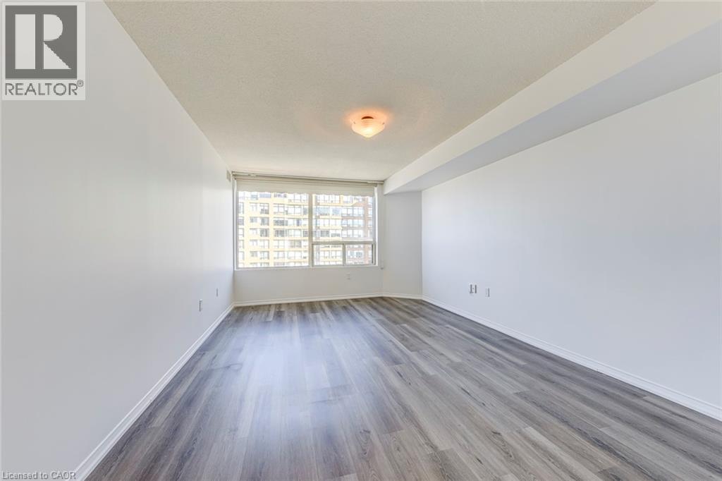 Spacious interior featuring wood-finish flooring, a large window, white walls, and a ceiling-mounted light fixture - 1270 Maple Crossing Boulevard Unit# 1705, Burlington, ON - Indoor Photo Showing Other Room