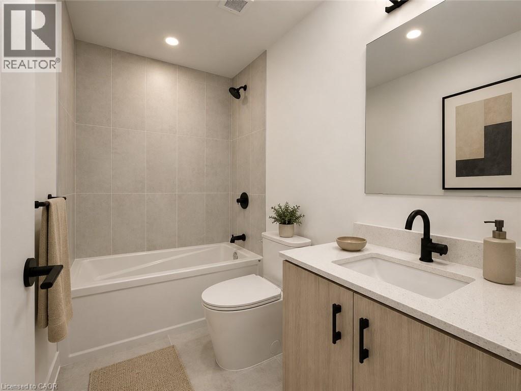 Bathroom featuring a light wood-finish vanity with black hardware, a light-colored solid surface countertop with an integrated sink, a sleek black faucet, a large frameless mirror, and a white bathtub - 4878 Powers Common Unit# C-1108, Burlington, ON - Indoor Photo Showing Bathroom