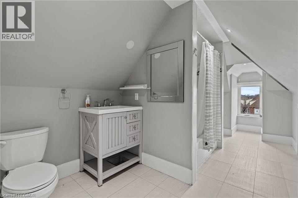 Bathroom featuring sloped ceilings, a single vanity with a solid surface countertop, a wall-mounted mirror, white ceramic tile flooring, and a shower with a white tile surround - 181 West Avenue N Unit# 4, Hamilton, ON - Indoor Photo Showing Bathroom