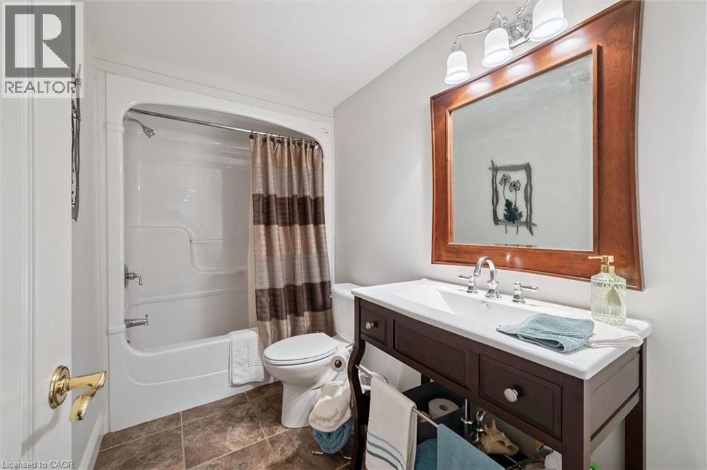 Bathroom featuring a white bathtub and shower unit, a dark wood-finish vanity with a white countertop and integrated sink, a large wood-framed mirror, and a three-light vanity fixture - 20 Bonniewood Drive, Drayton, ON - Indoor Photo Showing Bathroom