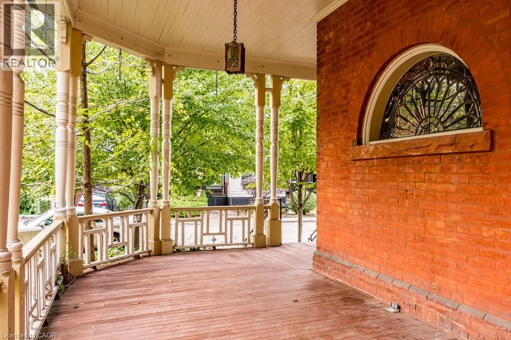 Expansive covered porch featuring wood flooring, decorative railing, turned columns, a ceiling-mounted lantern fixture, and a prominent arched window with intricate metalwork - 95-97 S Fairholt Road S, Hamilton, ON - Outdoor With Deck Patio Veranda With Exterior