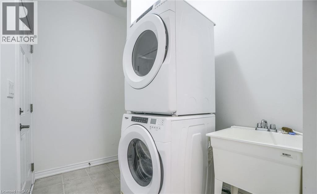 Laundry area featuring a stackable washer and dryer, a utility sink with a chrome faucet, white walls, and light-toned floor tiles - 14 Faye Street, Brampton, ON - Indoor Photo Showing Laundry Room