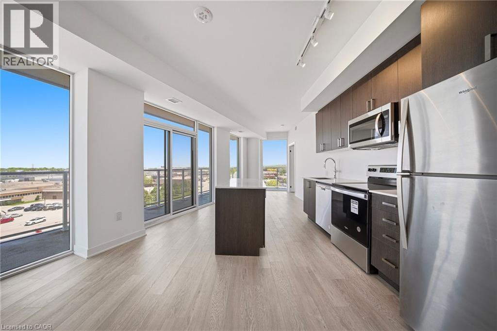 Open-concept living space featuring wood-finish flooring, a contemporary kitchen island with white countertop, dark wood-finish cabinetry, and stainless steel appliances - 2782 Barton Street E Unit# 516, Hamilton, ON - Indoor Photo Showing Kitchen