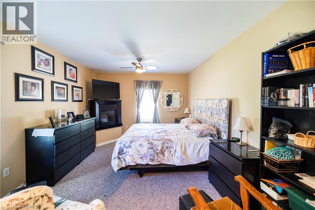 Carpeted bedroom featuring a corner fireplace, ceiling fan with integrated lighting, and a window with drapes - 100 Mechanic Street W, Waterford, ON - Indoor Photo Showing Bedroom With Fireplace