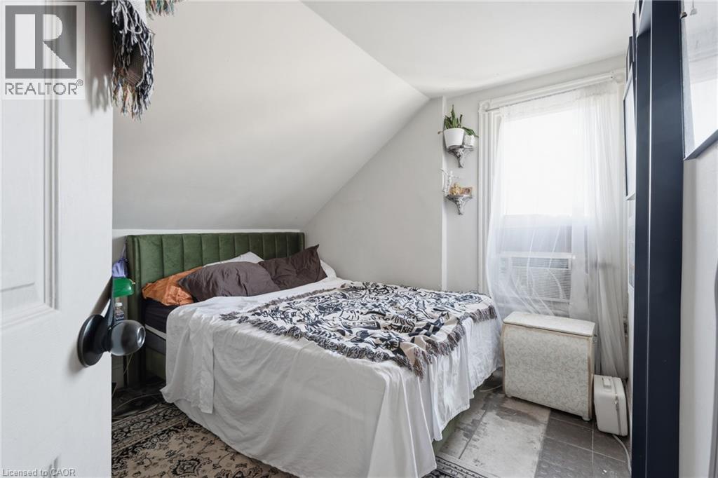 Sloped ceiling room featuring a window with sheer curtains, a dark-framed built-in element, and patterned area rugs over a tiled floor - 355 Barton Street E, Hamilton, ON - Indoor Photo Showing Bedroom