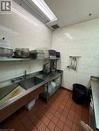 Commercial-grade stainless steel sink and countertop space with wall-mounted shelving, white tiled walls, and terracotta-toned floor tiling -