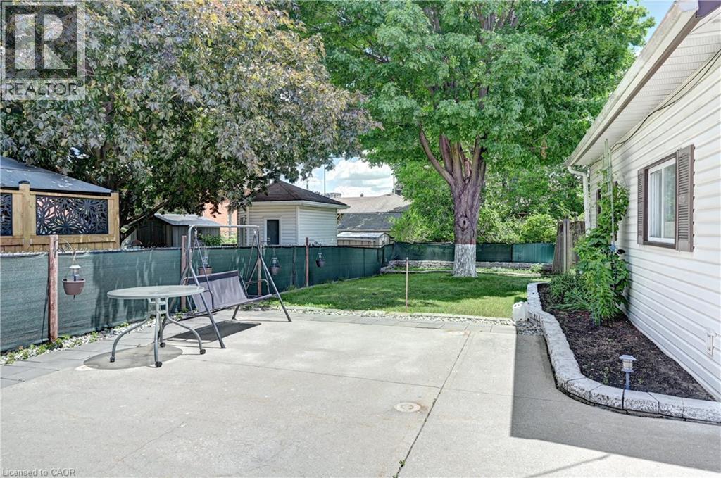 Spacious backyard with expansive concrete patio, lush green lawn, mature trees, and a landscaped garden bed with stone edging - 158 Plymouth Road, Kitchener, ON