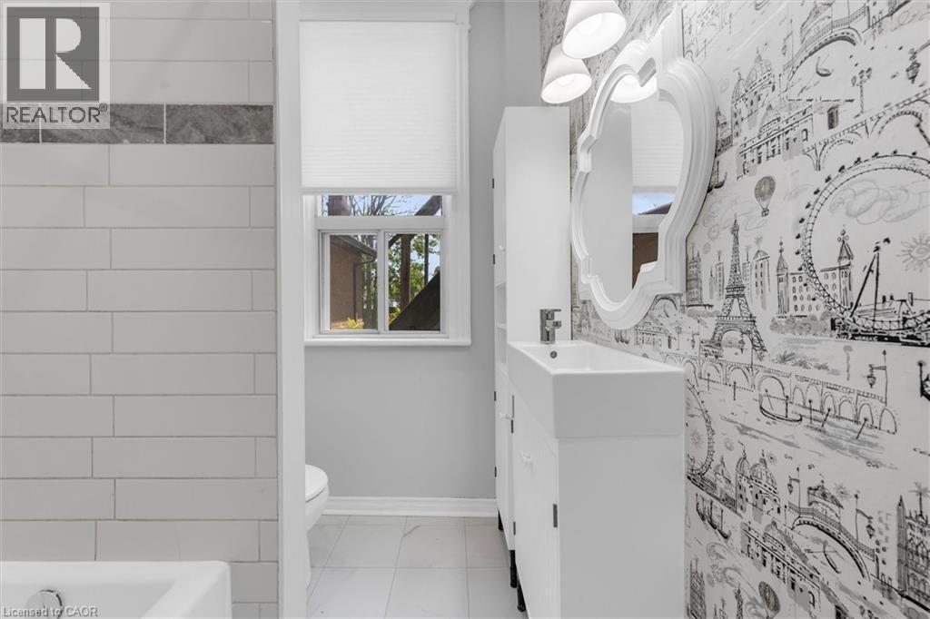 Bathroom featuring white subway tile, a decorative accent tile band, white vanity with integrated sink, contemporary wallpaper, and a wall-mounted mirror with overhead lighting - 205 Ferguson Avenue S, Hamilton, ON - Indoor Photo Showing Bathroom