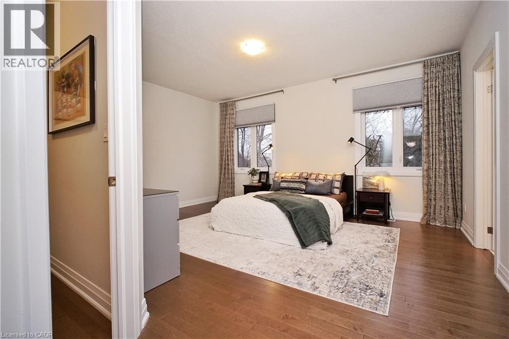 Bedroom featuring wood-finish flooring, two windows with roller shades and drapery, overhead lighting, and white baseboards - 63 Ian Ormston Drive, Kitchener, ON - Indoor