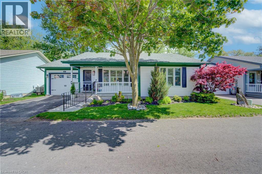 White siding exterior with a dark green roof trim, featuring a covered porch with white railings, a front-facing attached garage, and a manicured lawn with mature trees and shrubbery - 137 Wesley Crescent, Waterloo, ON