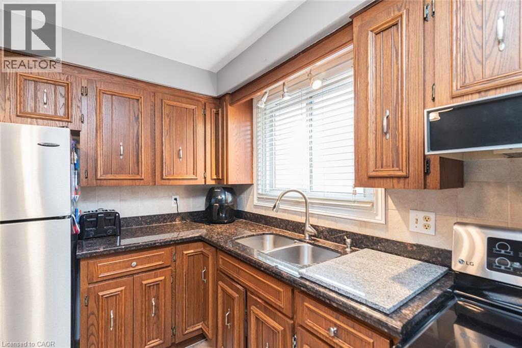 Kitchen featuring wood cabinetry, dark-toned countertops, a stainless steel double-basin sink with a gooseneck faucet, and a tiled backsplash - 366 Berkindale Drive, Hamilton, ON