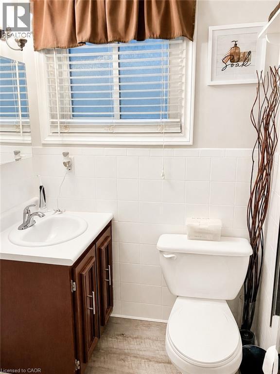 Bathroom featuring a window with white blinds, white wall tiling, a white toilet, and wood-finish flooring - 366 Berkindale Drive, Hamilton, ON