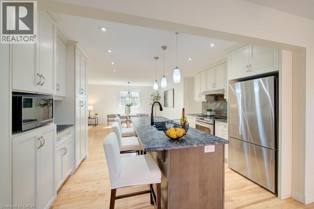 Modern kitchen featuring a central island with a dark countertop and pendant lighting, stainless steel appliances, extensive white cabinetry, and wood-finish flooring - 60 Mayfield Avenue, Waterloo, ON