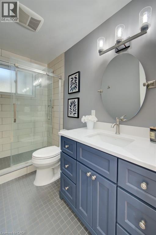 Bathroom featuring a navy blue vanity with a white countertop and integrated sink, a glass-enclosed shower with subway tile surround, an oval mirror, and a three-light vanity fixture - 60 Mayfield Avenue, Waterloo, ON