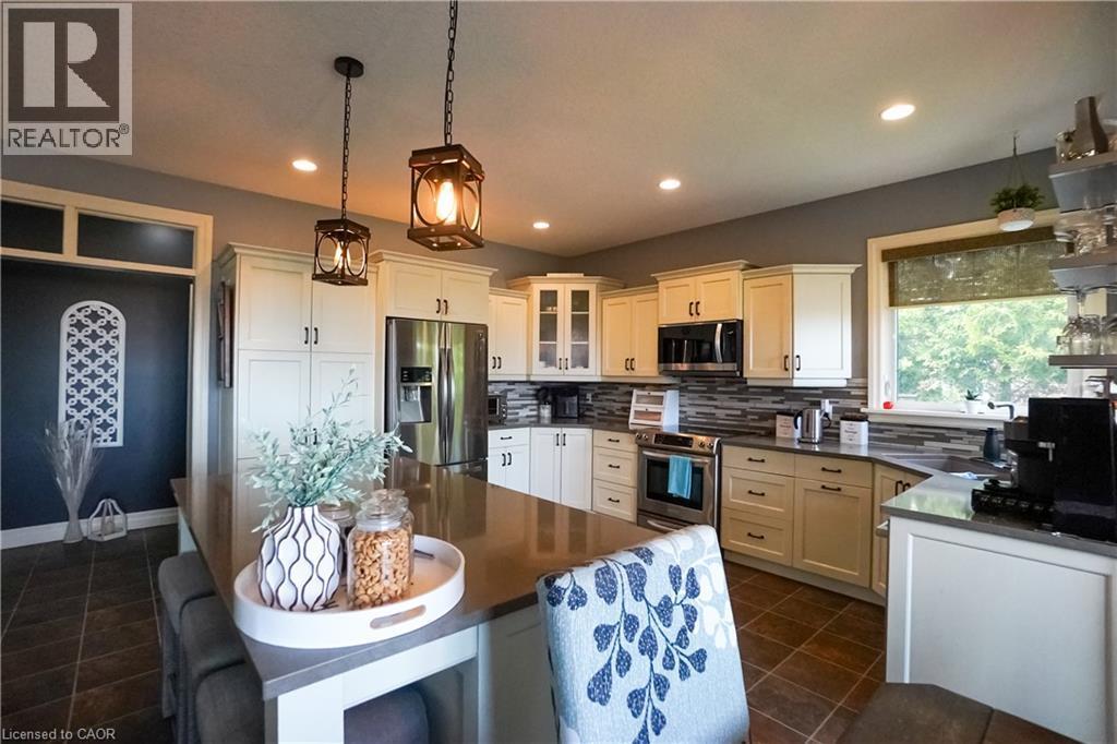 Kitchen featuring a large central island with a solid surface countertop, cream-colored cabinetry, stainless steel appliances, a multi-tone tile backsplash, and dark tile flooring - 365 Poldon Drive, Norwich, ON