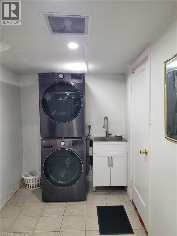 Laundry area featuring stacked appliances, a utility sink with a tall faucet, white base cabinetry, and neutral tile flooring - 153 Arnold Street, Kitchener, ON