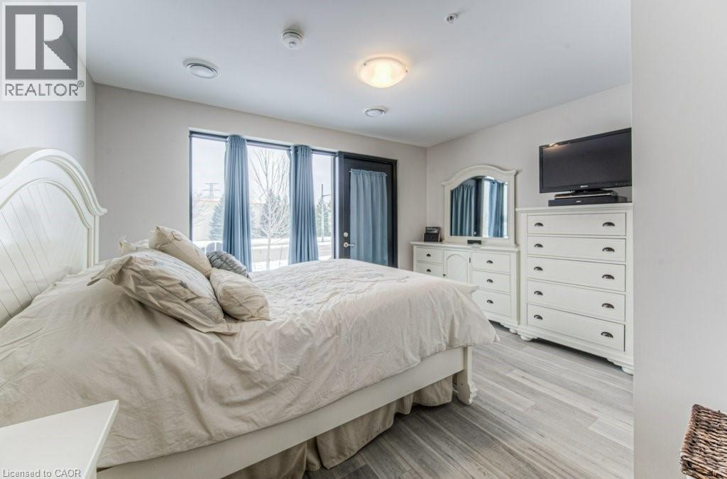 Bright bedroom featuring large windows, light wood-finish flooring, a contemporary ceiling light fixture, and recessed ceiling vents - 5 Wake Robin Drive Unit# 107, Kitchener, ON