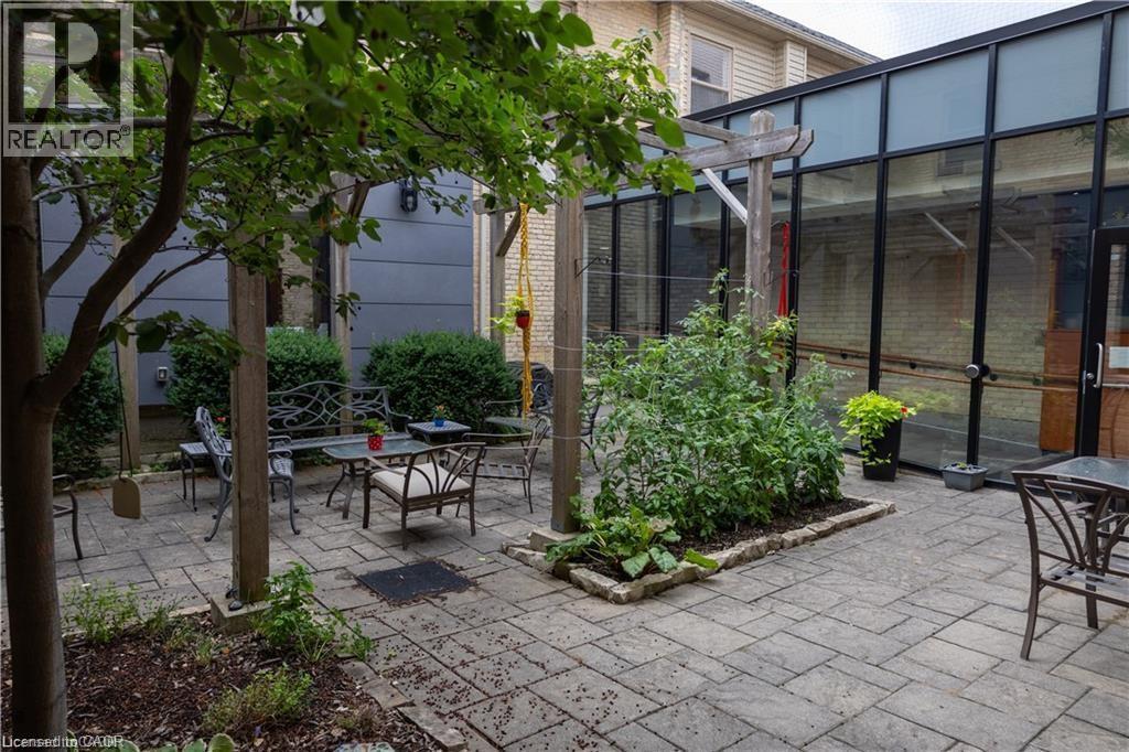 Paved patio area featuring an overhead pergola with mature greenery, a built-in garden bed, and a glass-enclosed structure - 365 Colborne Street, Brantford, ON