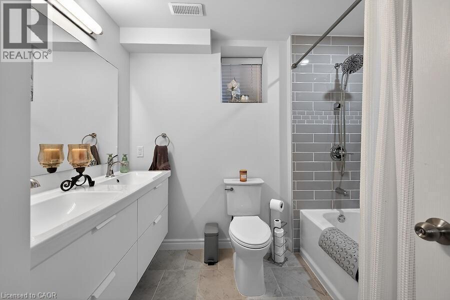 Bathroom featuring a dual-sink vanity with under-sink storage, a subway tile shower surround with an accent band, a white bathtub, and light-toned floor tiling - 109 Ray Street N, Hamilton, ON - Indoor Photo Showing Bathroom