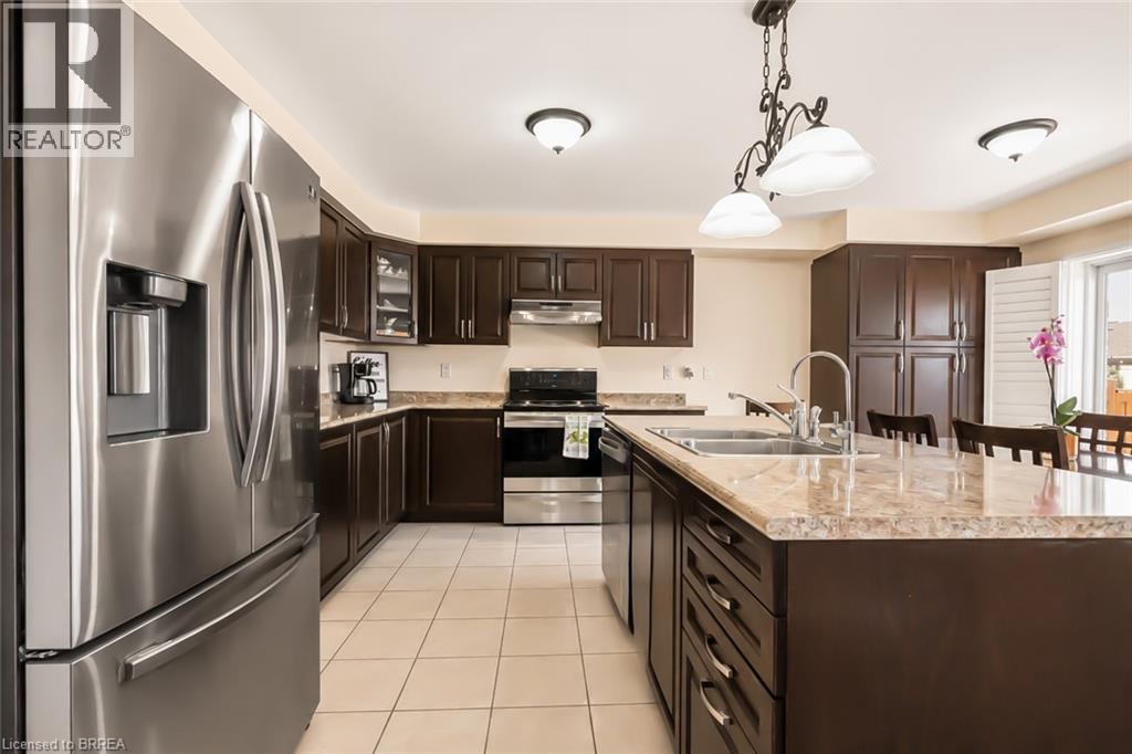 Kitchen featuring a large central island with a double basin sink, dark wood-finish cabinetry, stainless steel appliances, and light-toned tile flooring - 26 Pollard Street, Brantford, ON - Indoor Photo Showing Kitchen With Stainless Steel Kitchen With Double Sink