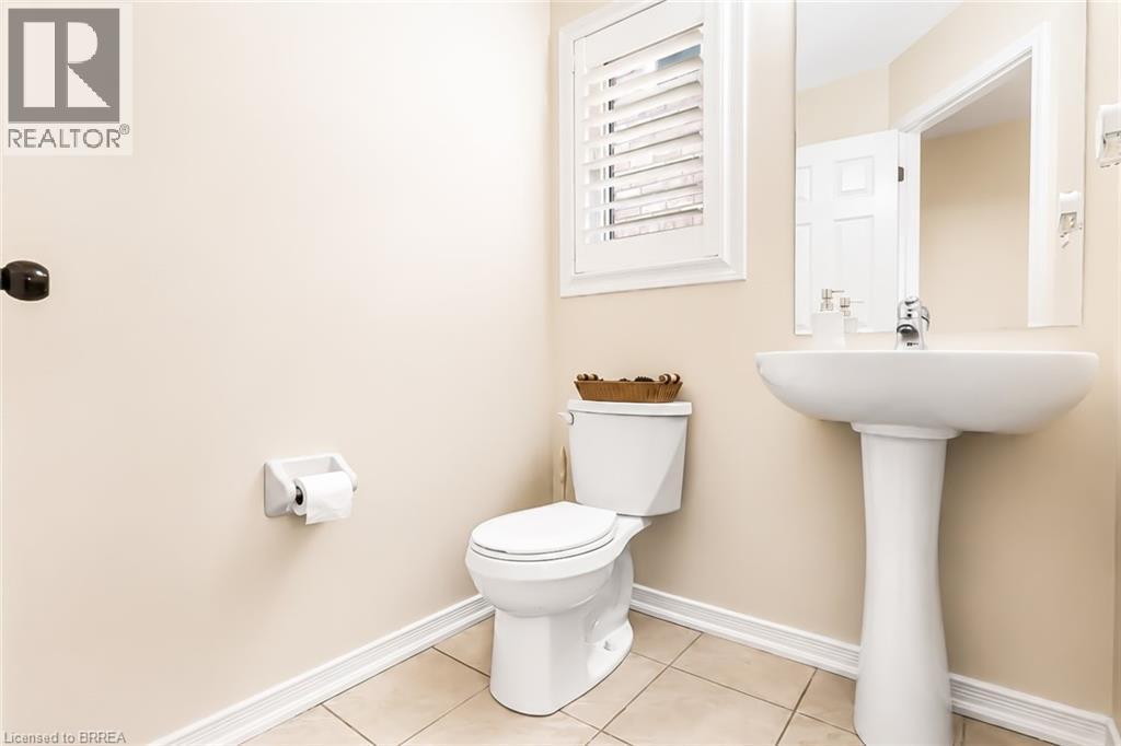 Pedestal sink with chrome faucet, ceramic toilet, window with white plantation shutters, wall-mounted mirror, and tile flooring - 26 Pollard Street, Brantford, ON - Indoor Photo Showing Bathroom