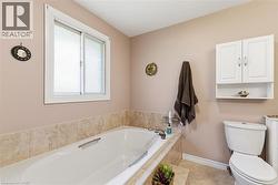 Soaking tub with a tiled surround, white sliding window, white wall-mounted storage cabinet with shelving, and tile flooring -
