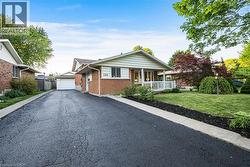 Brick exterior residence featuring a covered front porch with white railings, a front lawn with mature trees and shrubbery, and a paved driveway extending to a detached garage -