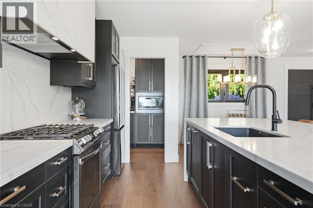 Contemporary kitchen featuring dark cabinetry, light countertops, wood-finish flooring, a kitchen island with an integrated sink and black faucet, and a large window with exterior greenery - 71 Miles Road, Hamilton, ON