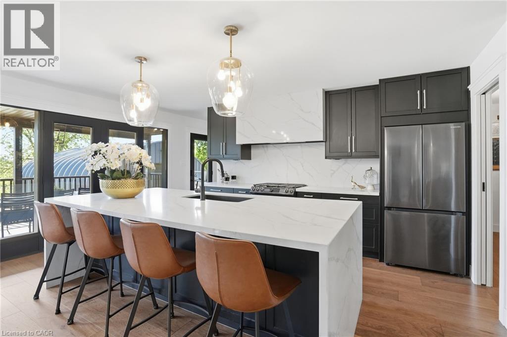 Modern kitchen featuring a large waterfall island with integrated sink, black cabinetry, stainless steel appliances, and wood-finish flooring - 71 Miles Road, Hamilton, ON