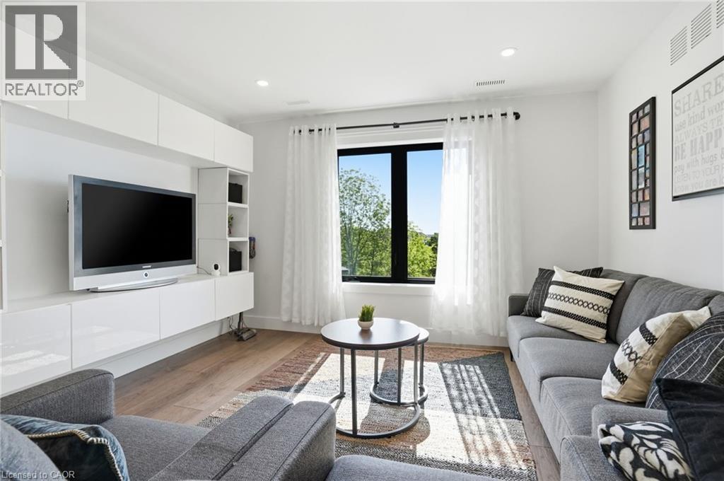 Light-filled living area featuring wood-finish flooring, a contemporary built-in media console with glossy white cabinetry, and a prominent window with dark framing - 71 Miles Road, Hamilton, ON