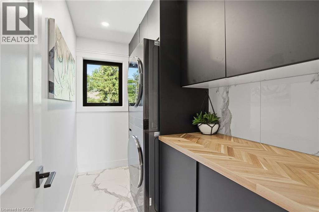 Contemporary utility space featuring stacked laundry appliances, rich wood-finish herringbone countertop, matte black cabinetry, and a white marble-patterned backsplash - 71 Miles Road, Hamilton, ON