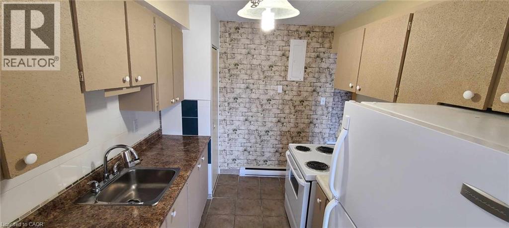 Galley kitchen featuring light brown cabinetry, speckled countertops, a stainless steel sink with a chrome faucet, white appliances, and a brick-patterned accent wall - 105 Conroy Crescent Unit# 505, Guelph, ON