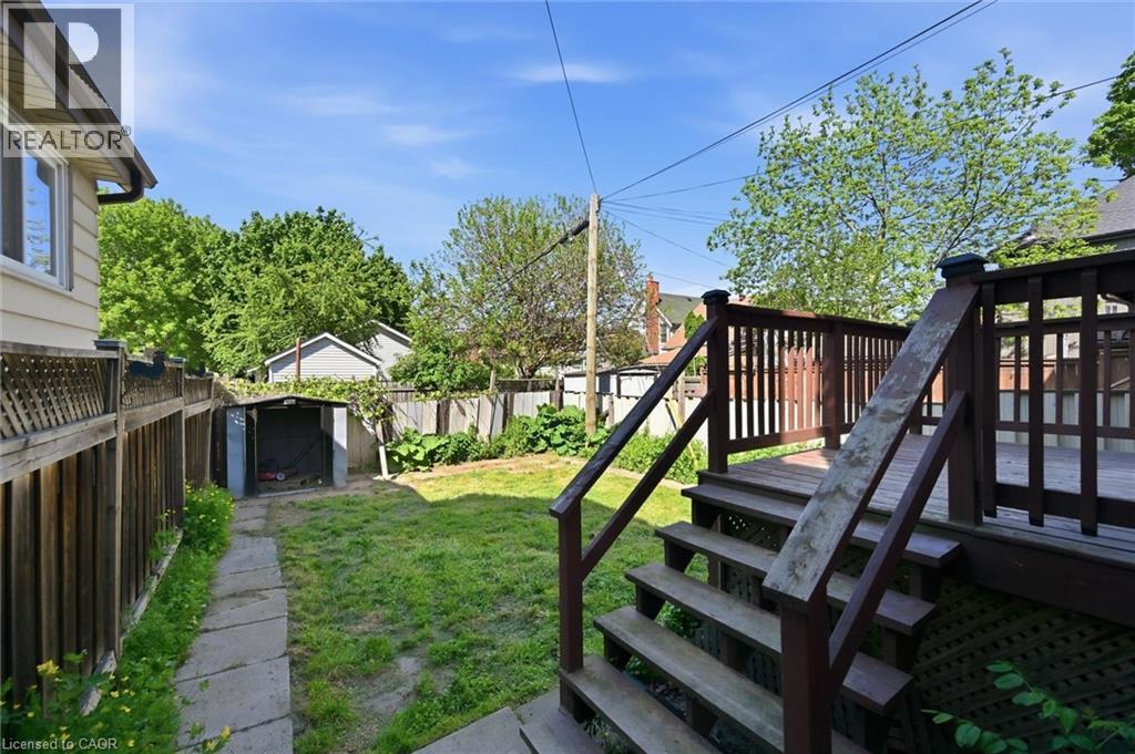Rear fully fenced yard featuring a wooden deck with railing and stairs, a grass lawn, a stone pathway, and a metal storage shed - 343 Cumberland Avenue, Hamilton, ON