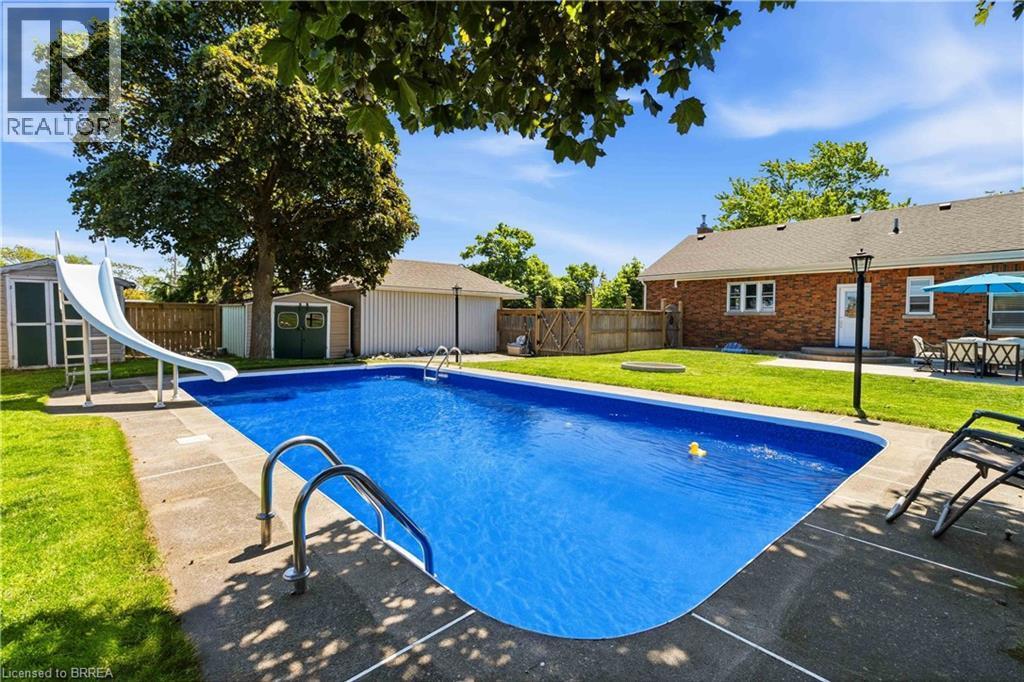 Expansive backyard featuring a rectangular in-ground swimming pool with concrete surround, a white pool slide, and two storage sheds - 134 Queensway Drive, Brantford, ON